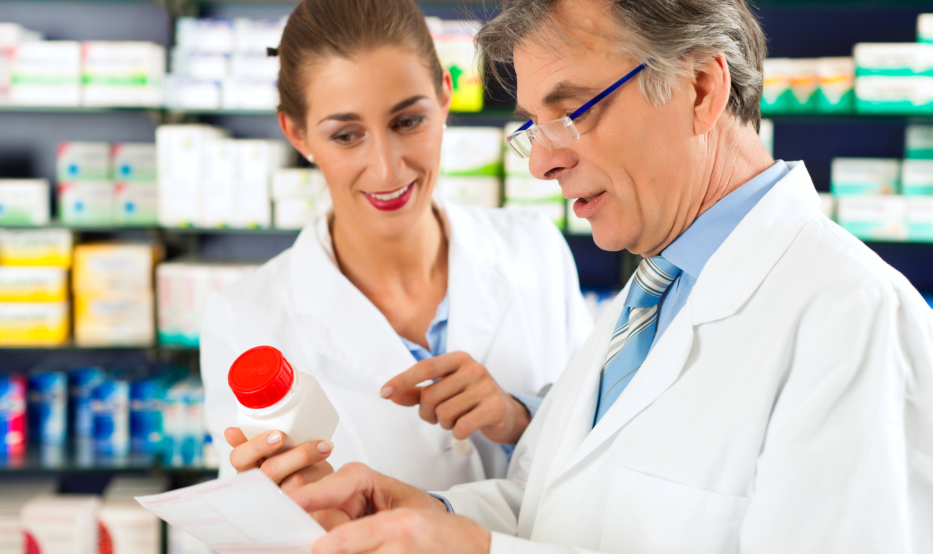 An older male pharmacist educating a younger female pharmacist while holding up a pill bottle