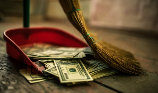 Someone using a broom to sweep dollar bills into a dust pan